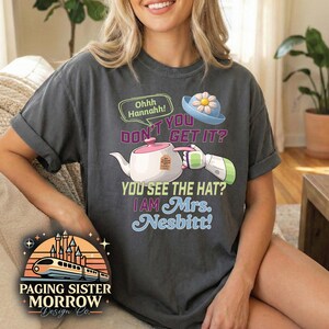 May include: Dark gray t-shirt with the text "DON'T YOU GET IT? YOU SEE THE HAT? I AM Mrs. Nesbitt!" and a cartoon image of a teapot, hat, and arm. The shirt also features a logo with a train and castle.