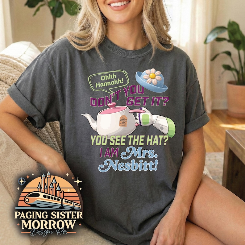 May include: Dark gray t-shirt with the text "DON'T YOU GET IT? YOU SEE THE HAT? I AM Mrs. Nesbitt!" and a cartoon image of a teapot, hat, and arm. The shirt also features a logo with a train and castle.