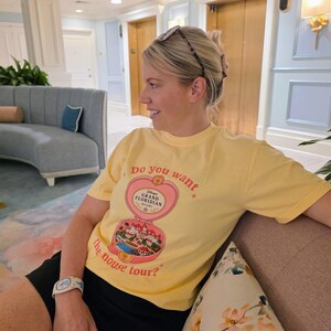 Puede incluir: Una camiseta de manga corta de color amarillo p&aacute;lido con un gr&aacute;fico de un relicario en forma de coraz&oacute;n que se abre para revelar una escena del Disney's Grand Floridian Resort. El texto dice "do you want the mouse tour?" con estrellas.
