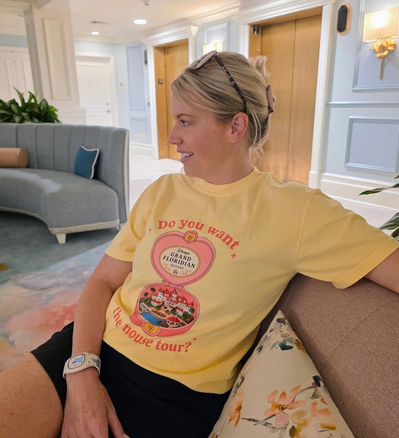 Puede incluir: Una camiseta de manga corta de color amarillo p&aacute;lido con un gr&aacute;fico de un relicario en forma de coraz&oacute;n que se abre para revelar una escena del Disney's Grand Floridian Resort. El texto dice "do you want the mouse tour?" con estrellas.