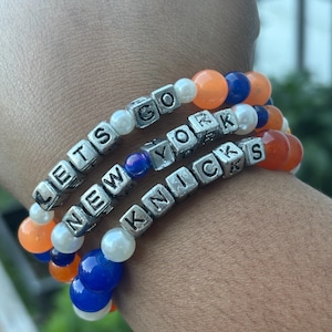 Peut inclure: Trois bracelets de perles empilés composés de perles rondes orange, bleues et blanches. Les bracelets intègrent des perles carrées argentées avec des lettres formant la phrase LETS GO NEW YORK KNICKS répartie sur les trois rangs.