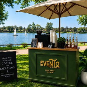 May include: A green mobile coffee bar with a beige umbrella, featuring the text EVENTO POP UP BARS.CO SOUTH AFRICA ESTD 2024. A black chalkboard sign reads Good days start with COFFEE and LAKE VIEWS HOMESTEAD LAKE BENONI, with coffee supplies.