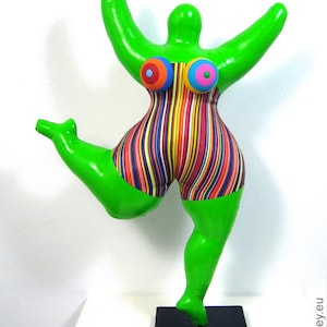 Könnte beinhalten: Eine skurrile Skulptur einer grünen Figur in Tanzpose. Die Figur trägt einen gestreiften Badeanzug in leuchtenden Farben und zwei runde, farbenfrohe brustähnliche Formen. Sie steht auf einem schwarzen quadratischen Sockel, die Arme erhoben und ein Bein ausgestreckt.