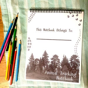 Puede incluir: Un cuaderno de seguimiento de animales con un diseño de silueta de bosque. El cuaderno tiene una encuadernación en espiral e incluye el texto "This Notebook Belongs To:" y "Animal Tracking Notebook." Una colección de lápices de colores de varios colores está dispuesta a la izquierda.