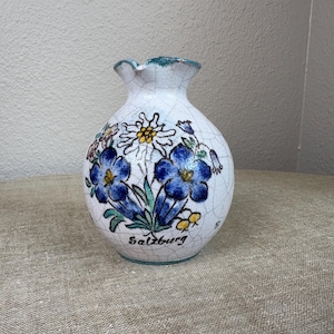 May include: A white ceramic vase with a cracked glaze, featuring blue floral designs and the word "Salzburg" in black. The vase has a ruffled rim with green trim and is likely a decorative item.