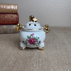 May include: A white porcelain potpourri jar with a lid, featuring a floral pattern of pink roses and green leaves. The jar has gold-painted handles, feet, and a finial on the lid. The lid is perforated with small circular holes for scent release.