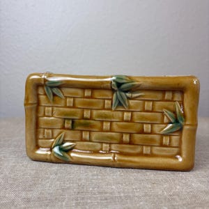 May include: A rectangular ceramic container with a woven basket design in shades of tan and brown. Green bamboo leaves are placed on the corners. The container has a glossy finish.