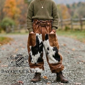 Puede incluir: Pantalones de cuero marrón con paneles de piel de vaca, cinturón ancho y bolsillos. Los pantalones se llevan con botas marrones y un suéter verde. La imagen incluye el texto "Bavaria Craft" y "Handcrafted Leather Goods".