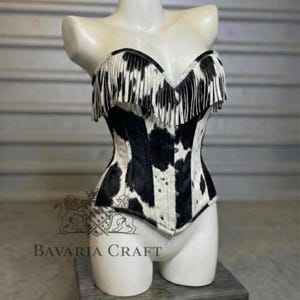 May include: A strapless corset top featuring a black and white cow print pattern with long fringe detailing along the neckline. The garment includes a front closure with visible metal buttons and is displayed on a white mannequin torso.