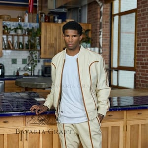 May include: Cream-colored bomber jacket and matching pants with brown trim. The jacket has a zipper closure and ribbed cuffs. The outfit is paired with a white t-shirt. The text "Bavaria Craft" is visible on the counter.