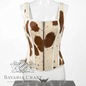 May include: A sleeveless corset top featuring a brown and white cowhide pattern with a textured hair-on-hide finish. The garment has a square neckline, a central vertical metal zipper closure, and white piping along the seams for structure.