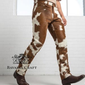 Op de afbeelding: Bruin-witte koeienhuid broek. De broek heeft een klassiek vijf-zakken ontwerp en een rechte pijp. Het koeienhuidpatroon is een mix van bruine en witte vlekken. De broek wordt gedragen met een bruine riem en bruine leren laarzen. Het merk "BAVARIA CRAFT" is zichtbaar.