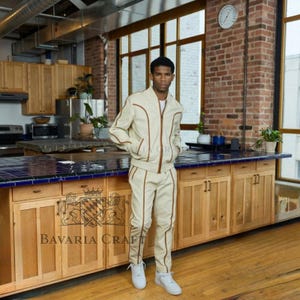 May include: A beige tracksuit with brown trim, consisting of a zip-up jacket and matching pants. The outfit is worn with white sneakers. The text "Bavaria Craft" is visible on the wooden cabinet.