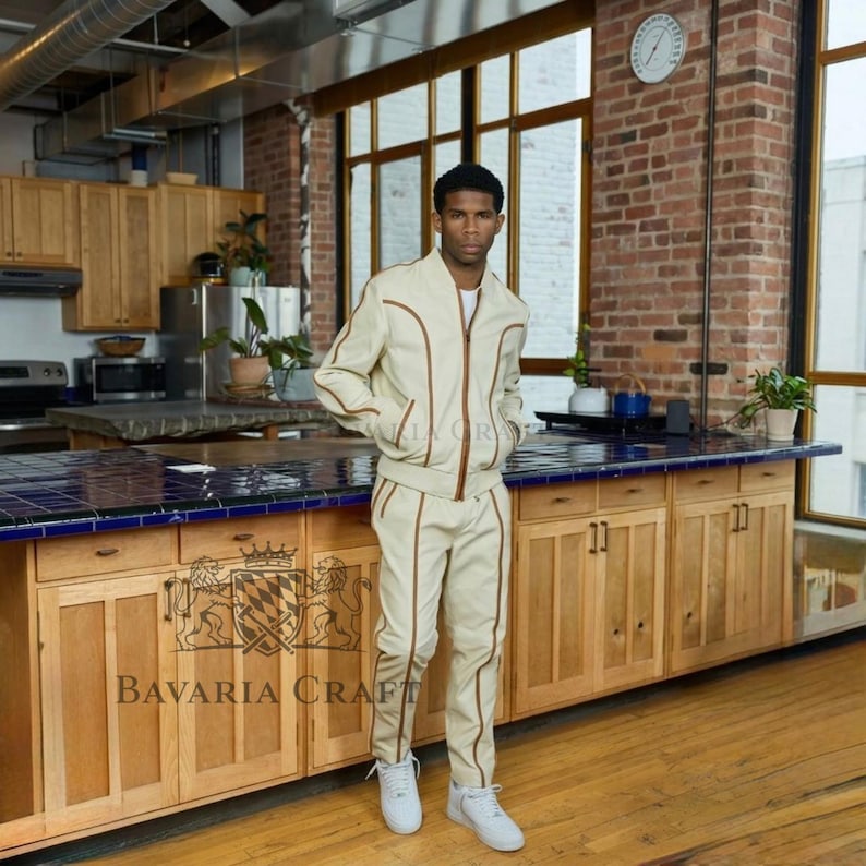 May include: A beige tracksuit with brown trim, consisting of a zip-up jacket and matching pants. The outfit is worn with white sneakers. The text "Bavaria Craft" is visible on the wooden cabinet.
