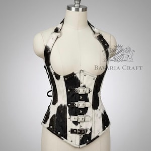 May include: A halter-style corset featuring a black and white cow print pattern. The garment includes four horizontal silver-toned metal buckles on the front, silver rivets along the seams, and a halter neck strap secured with metal rings.
