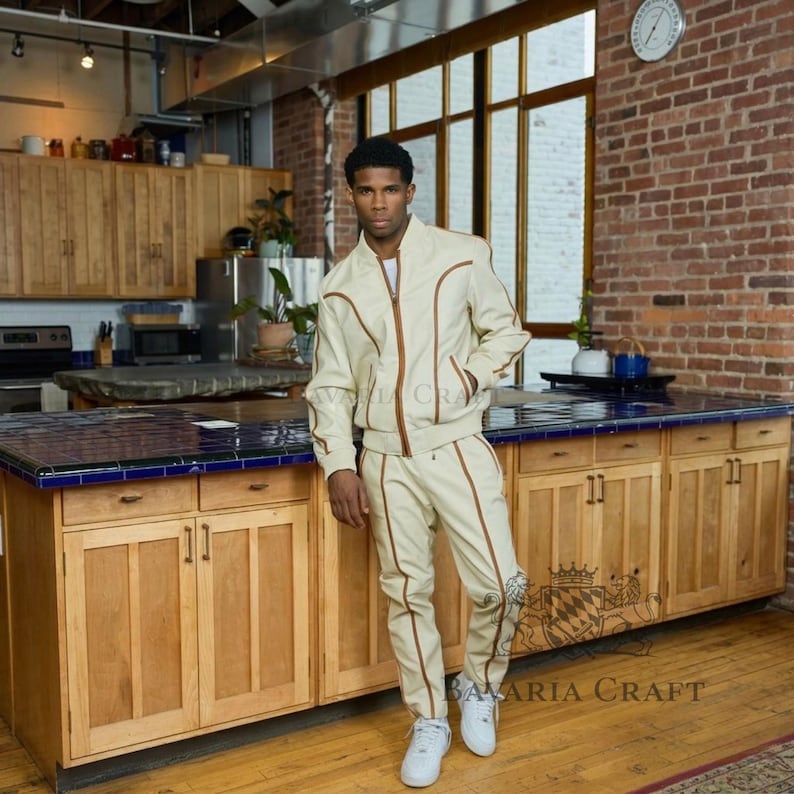 May include: Cream-colored track suit with brown trim. The jacket has a zipper closure and the pants have a drawstring. The suit is worn with white sneakers. The logo "BAVARIA CRAFT" is visible on the pants.