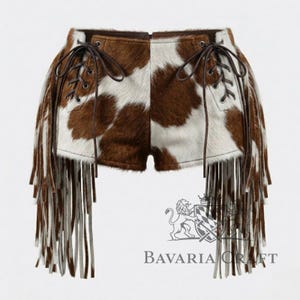 Puede incluir: Pantalones cortos de piel de vaca con un estampado de manchas marrones y blancas. Los pantalones cortos tienen cordones de cuero marrón y largas flecos en los laterales. La marca "Bavaria Craft" está impresa en la parte inferior.