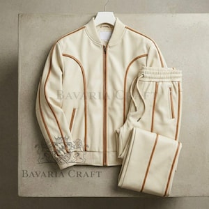 May include: Cream-colored leather track suit set, including a zip-up bomber jacket and matching pants. The jacket and pants feature brown trim and the text "BAVARIA CRAFT". The jacket is hanging on a white hanger.