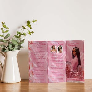 May include: A pink memorial service program, featuring the text "Order of Service," "Forever Loved," and "In Loving Memory." The program includes photos and text, with a floral design. The program is displayed on a wooden surface.