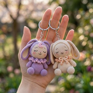 May include: Two crochet bunny keychains, one in lavender and one in cream, each featuring a small ruffled collar, closed eyes, and a crescent moon detail on the forehead or ear. Both plush figures are attached to silver metal key rings.