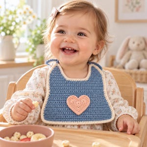 Pode incluir: Um babador de crochê azul para bebê com um coração aplicado na cor pêssego ao centro e uma borda na cor creme. O babador é feito com um padrão de ponto denso e inclui laços para amarrar na parte superior, sendo um acessório funcional.