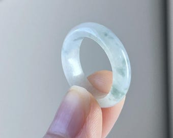 May include: A translucent jade ring with a pale green hue and subtle variations in color. The ring is held up by a person's fingers, showcasing its smooth, polished surface. The ring's design is simple and elegant, suitable for everyday wear.