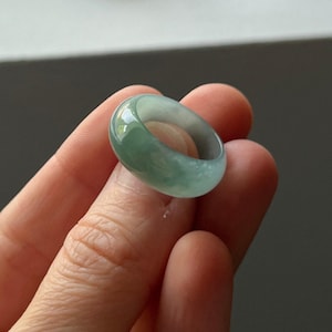 Op de afbeelding: Een doorschijnende jade ring met een glad, gepolijst oppervlak. De ring heeft een mix van lichtgroene en witte tinten, waardoor een gemarmerd effect ontstaat. De ring is een effen band, die de natuurlijke variaties in de steen laat zien.