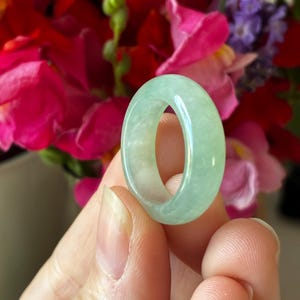 Könnte beinhalten: Ein hellgrüner Jade-Ring. Der Ring ist ein massives Band mit einer glatten, polierten Oberfläche. Der Jade hat eine durchscheinende Qualität, und die Farbe ist ein weiches, helles Grün. Der Hintergrund zeigt eine verschwommene Anordnung bunter Blumen.
