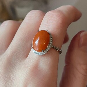 Op de afbeelding: Ovale oranje edelsteenring met een halo van kleine, heldere stenen, gezet in een zilverkleurige band. De edelsteen van de ring heeft een levendige, warme oranje kleur. De ring wordt op een vinger getoond.
