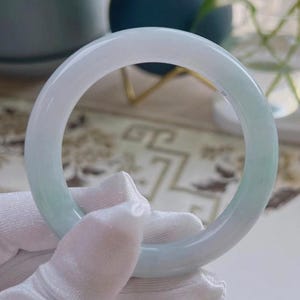 May include: A pale green and white jade bangle bracelet. The bracelet has a smooth, polished surface. The jade is held by a gloved hand. The bangle is a circular shape.
