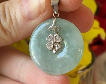 Op de afbeelding: Een ronde, lichtgroene jade hanger met een zilveren beugel. De hanger heeft een zilveren klavertjevier ontwerp met kleine, heldere stenen. De jade heeft een glad, gepolijst oppervlak en is licht doorschijnend. De hanger wordt vastgehouden.