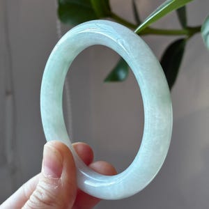 May include: A smooth, translucent pale green jade bangle bracelet with a polished finish. The circular jewelry piece features subtle white cloudy inclusions and a rounded profile, showcasing a natural light green color throughout the stone.