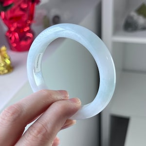 Könnte beinhalten: Ein hellblaues Jade-Armreif-Armband. Die glatte, polierte Oberfläche des Armbands reflektiert das Licht und zeigt seine durchscheinende Qualität. Das Armband wird in einer Hand gehalten, mit einem Aufkleber auf der Innenseite. Der Hintergrund ist unscharf.