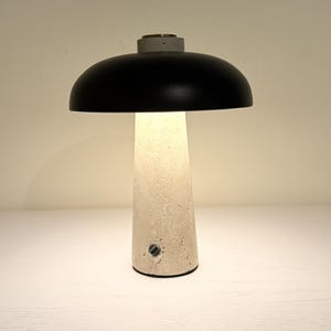 May include: A modern table lamp with a black dome-shaped shade and a textured, off-white concrete base. The lamp emits a warm glow, and the base has a small button. The lamp is a stylish addition to any home decor.