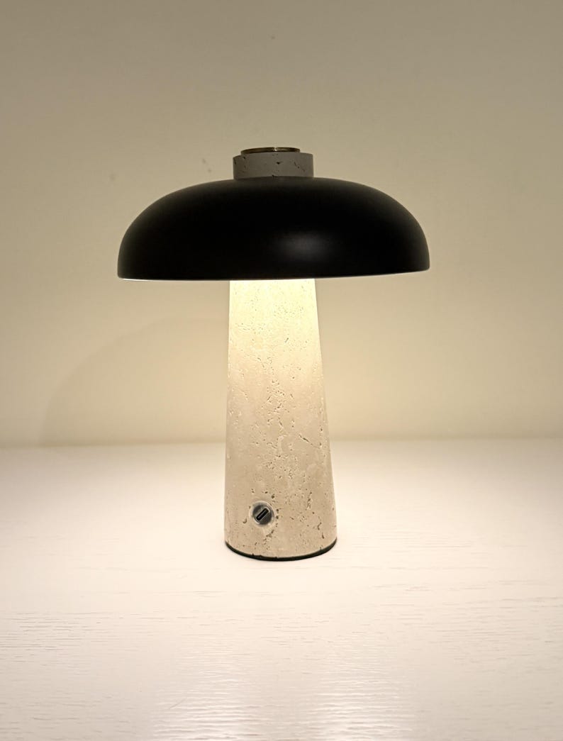 May include: A modern table lamp with a black dome-shaped shade and a textured, off-white concrete base. The lamp emits a warm glow, and the base has a small button. The lamp is a stylish addition to any home decor.
