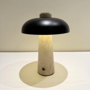 May include: A modern table lamp with a black lampshade and a beige, cone-shaped base. The lamp is turned on, emitting a warm glow. The base has a textured, concrete-like appearance. The lamp casts a circular shadow on the white surface.