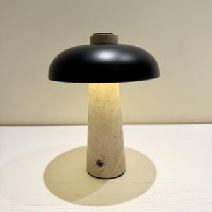 May include: A modern table lamp with a black, dome-shaped shade and a beige, conical base. The lamp is illuminated, casting a warm glow. The base appears to be made of stone or concrete. The lamp is on a white surface.