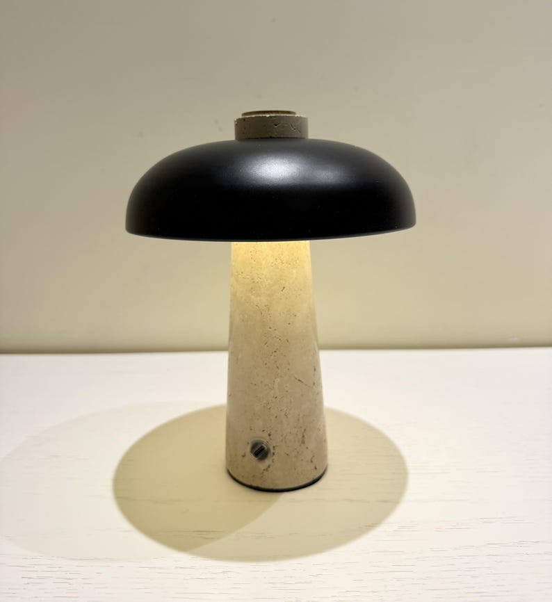 May include: A modern table lamp with a black, dome-shaped shade and a beige, conical base. The lamp is illuminated, casting a warm glow. The base appears to be made of stone or concrete. The lamp is on a white surface.