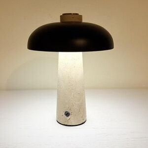 May include: A modern table lamp with a black dome-shaped shade and a textured, off-white base. The lamp emits a soft, warm light. The base has a cylindrical shape and a small power button. The top of the base has a small, circular, gold-colored accent.