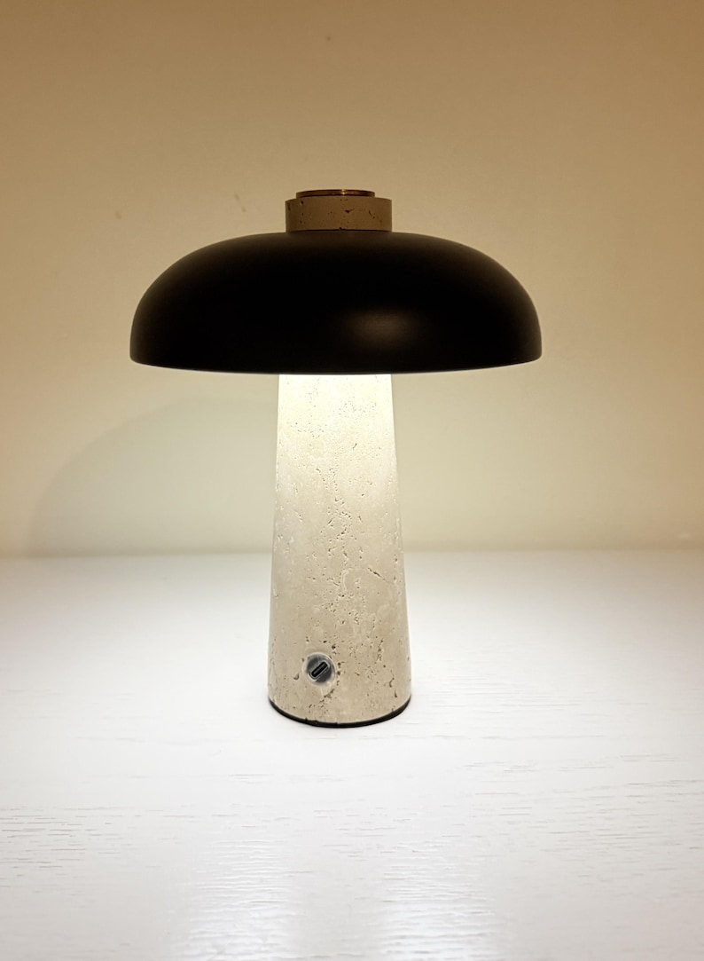 May include: A modern table lamp with a black dome-shaped shade and a textured, off-white base. The lamp emits a soft, warm light. The base has a cylindrical shape and a small power button. The top of the base has a small, circular, gold-colored accent.