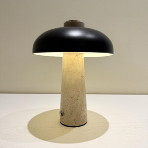 May include: A modern table lamp with a black dome-shaped shade and a beige, textured concrete base. The lamp emits a warm, inviting light. The lamp's design is minimalist and contemporary.