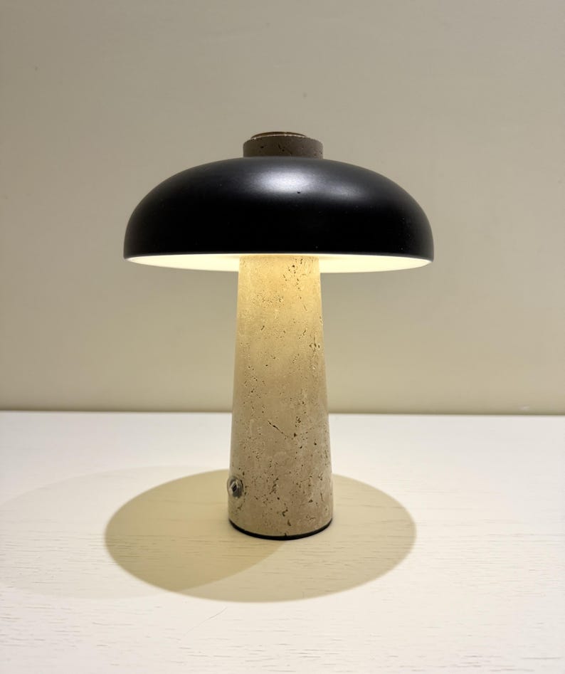 May include: A modern table lamp with a black dome-shaped shade and a beige, textured concrete base. The lamp emits a warm, inviting light. The lamp's design is minimalist and contemporary.