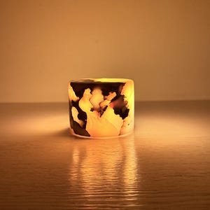 Puede incluir: Un portavelas cilíndrico con un diseño único. El portavelas presenta una mezcla de patrones negros, blancos y naranjas, creando un efecto marmolado. El portavelas está iluminado desde dentro, emitiendo un brillo cálido.