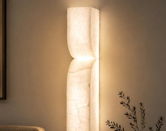 May include: A vertical wall sconce featuring a textured, translucent white stone-like material that emits a warm, soft glow. The fixture has a distinct split design in the center, creating two stacked, curved segments that illuminate the wall.