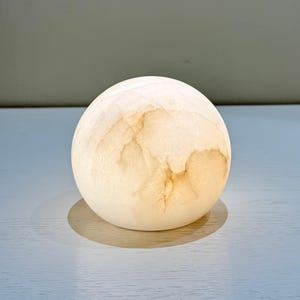 Puede incluir: Objeto decorativo esférico blanquecino con aspecto marmóreo y translúcido. El orbe presenta sutiles vetas marrones y se apoya sobre una superficie azul claro, proyectando una suave sombra. La estética general es minimalista y natural.