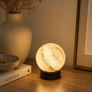 May include: A spherical lamp, reminiscent of the moon, with a warm glow, sitting on a dark base. A vase with dried flowers, a bowl, and a book titled "KINFOLK" are on a wooden surface. The lamp emits a soft light, creating shadows.