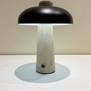 May include: A modern table lamp with a mushroom-shaped design. It features a black lampshade and a textured, light gray concrete base. The lamp is illuminated, casting a soft glow.