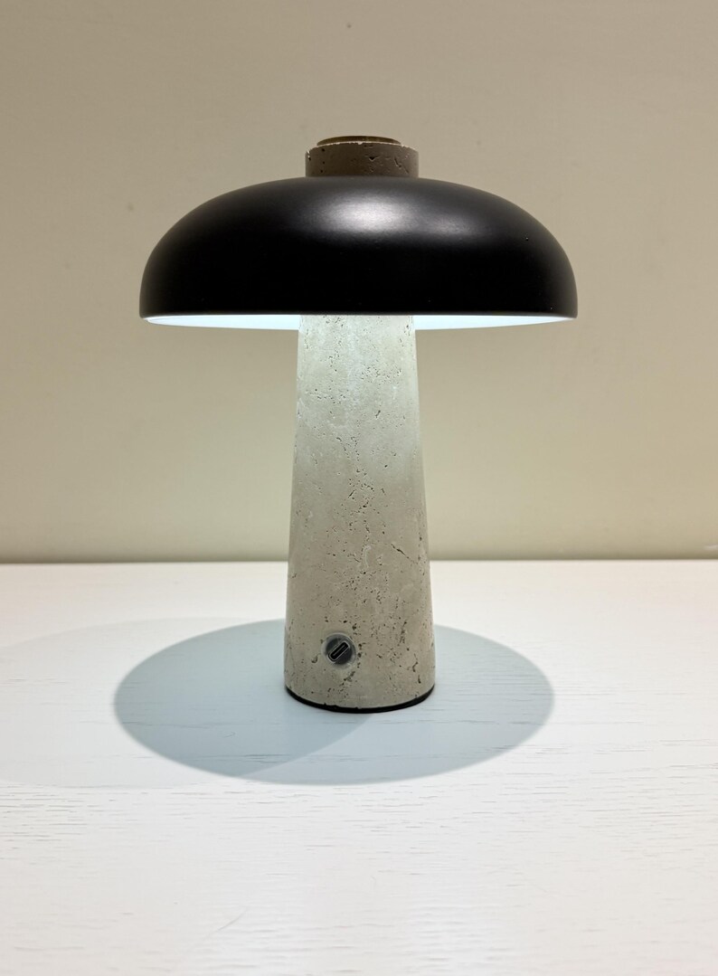 May include: A modern table lamp with a mushroom-shaped design. It features a black lampshade and a textured, light gray concrete base. The lamp is illuminated, casting a soft glow.
