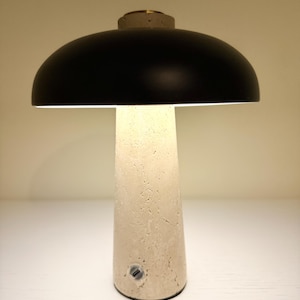 Puede incluir: Una lámpara de mesa moderna con una pantalla negra en forma de cúpula y una base texturizada de color blanquecino. La lámpara tiene una base cilíndrica y un pequeño botón de encendido circular. La parte superior de la lámpara tiene un pequeño acento circular dorado.