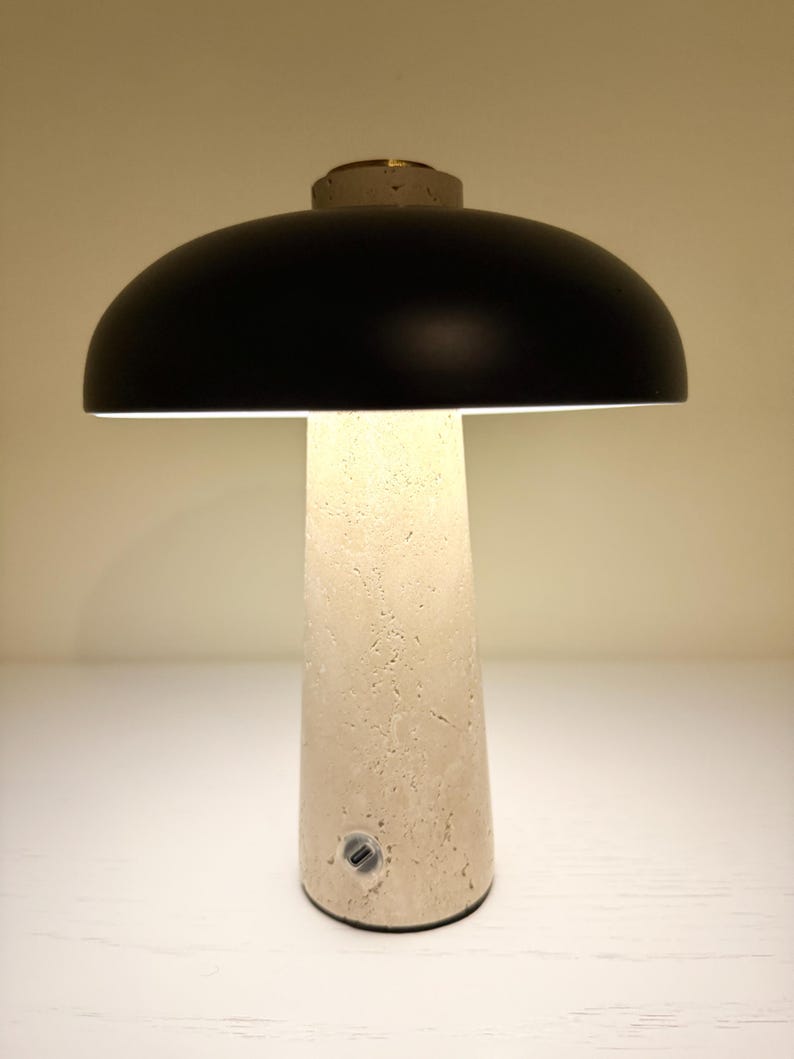May include: A modern table lamp with a black dome-shaped shade and a textured, off-white base. The lamp has a cylindrical base and a small, circular power button. The top of the lamp has a small, circular, gold-colored accent.